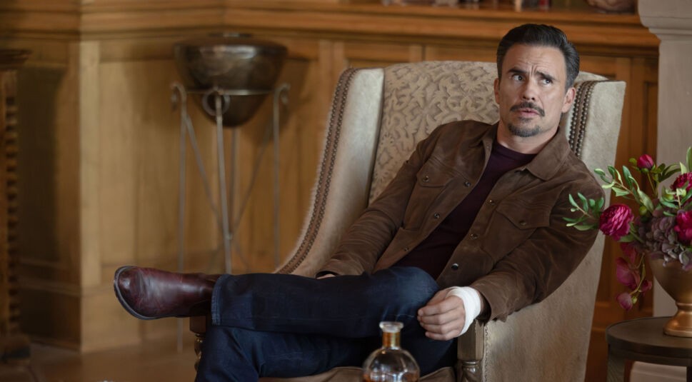 Auf dem Bild zur Dutton Ranch ist Juan Pablo Raba als Joaquin in einer stilvollen Innenraumszene zu sehen. Er sitzt entspannt in einem Sessel, trägt eine braune Jacke und blickt mit skeptischem, leicht angespanntem Ausdruck zur Seite. Die warme Beleuchtung und elegante Umgebung verleihen der Szene eine ruhige, aber unterschwellig gespannte Atmosphäre.