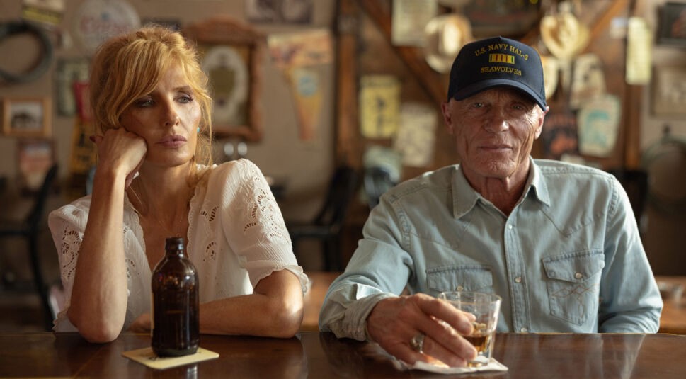 Auf dem Bild zur Dutton Ranch sitzen Kelly Reilly als Beth Dutton und Ed Harris als Everett McKinney an einer Bar in einer rustikalen Umgebung. Beth wirkt nachdenklich und stützt ihren Kopf auf die Hand, während Everett ruhig ein Glas in der Hand hält. Die Szene strahlt eine ruhige, leicht melancholische und zugleich spannungsgeladene Atmosphäre aus.