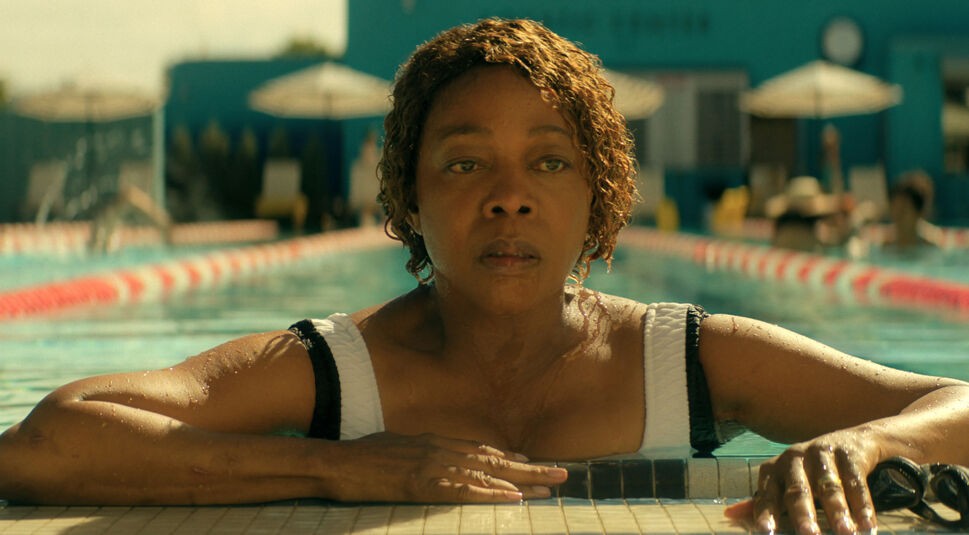 Auf dem Bild zu The Boroughs ist Judy, gespielt von Alfre Woodard, in einer ruhigen, aber nachdenklichen Szene in einem Außenpool zu sehen. Sie stützt sich mit beiden Armen am Beckenrand ab, während Wasser über ihre Schultern läuft. Ihr ernster, leicht erschöpfter Blick richtet sich geradeaus. Die helle Sonne und die ruhige Poolumgebung stehen im Kontrast zu ihrer angespannten und nachdenklichen Stimmung in The Boroughs.