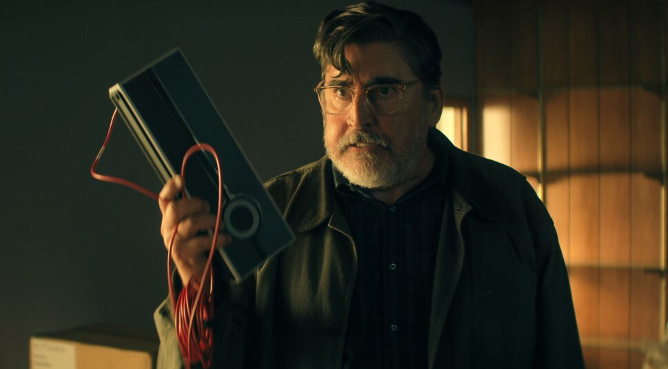 Auf dem Bild zu The Boroughs ist Sam, gespielt von Alfred Morina, in einer angespannten Szene in einem warm beleuchteten Raum zu sehen. Er trägt Brille und Bart und hält ein längliches technisches Gerät mit rotem Kabel in der Hand. Sein ernster Blick wirkt konzentriert und leicht besorgt, während er das Objekt prüfend anhebt.