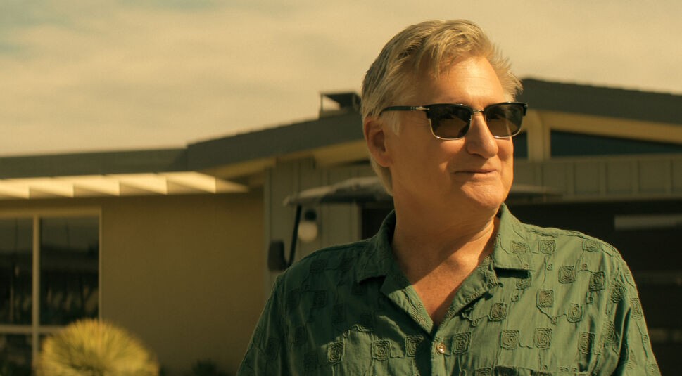 Auf dem Bild zu The Boroughs ist Jack, gespielt von Bill Pullman, in einer sonnigen Außenszene vor einem modernen Gebäude zu sehen. Er trägt eine dunkle Sonnenbrille und ein grünes, gemustertes Kurzarmhemd. Sein Gesichtsausdruck wirkt ruhig und selbstbewusst, während er leicht zur Seite blickt. Die warme Lichtstimmung verleiht der Szene eine entspannte, leicht geheimnisvolle Atmosphäre.