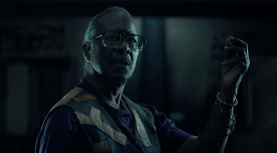 Auf dem Bild zu The Boroughs ist Art, gespielt von Clarke Peters, in einer düsteren Innenraumszene zu sehen. Er trägt eine Brille, ein dunkles Shirt und eine gemusterte Weste, während mehrere Armbänder sein Handgelenk schmücken. Mit erhobener Hand hält er ein kleines Objekt zwischen den Fingern und blickt ernst und nachdenklich zur Seite. Das kühle, schwache Licht verstärkt die geheimnisvolle und angespannte Stimmung der Szene aus The Boroughs.