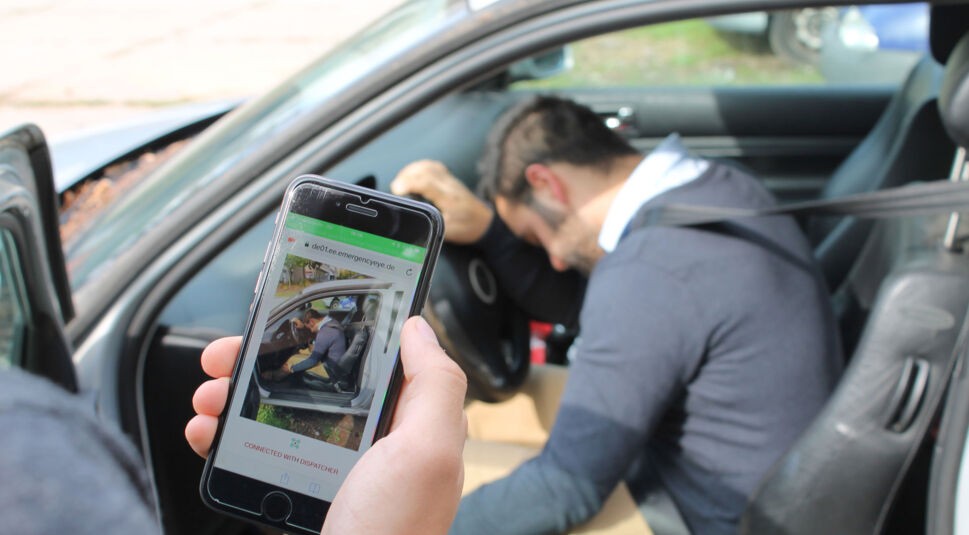 Ein Mann ruft mithilfe der Technologie EmergencyEye über sein Smartphone den Notruf, um einem bewusstlosen Autofahrer zu helfen.