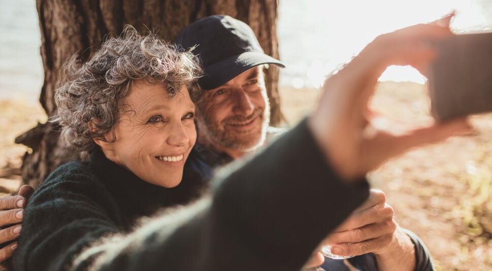 Happy senior couple taking their self portrait with their cell phone on camping day. Man and woman sitting together outdoors at campsite and taking selfie.