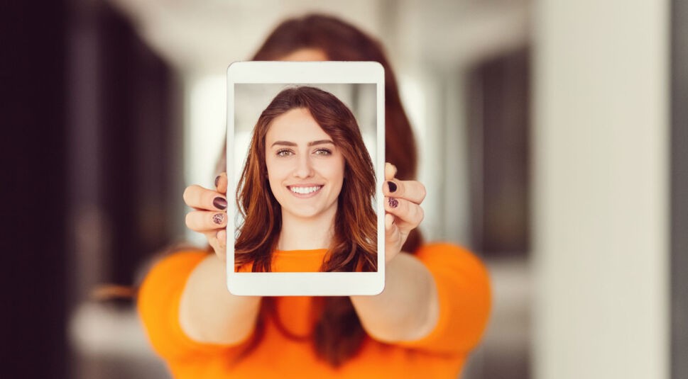 Frau hält eigenes Porträtfoto auf Tablet in die Kamera