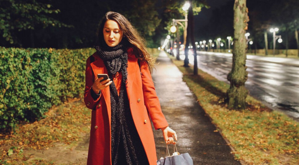 Frau im orangenen Mantel schaut nachts auf ihr Smartphone und trägt eine Einkaufstüte.