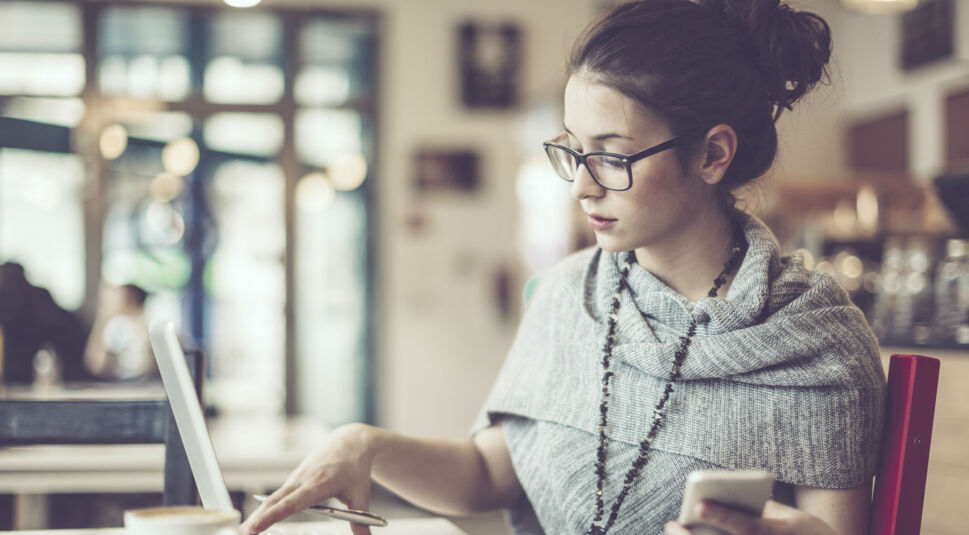 Frau arbeitet am Laptop und hält Smartphone in der Hand.