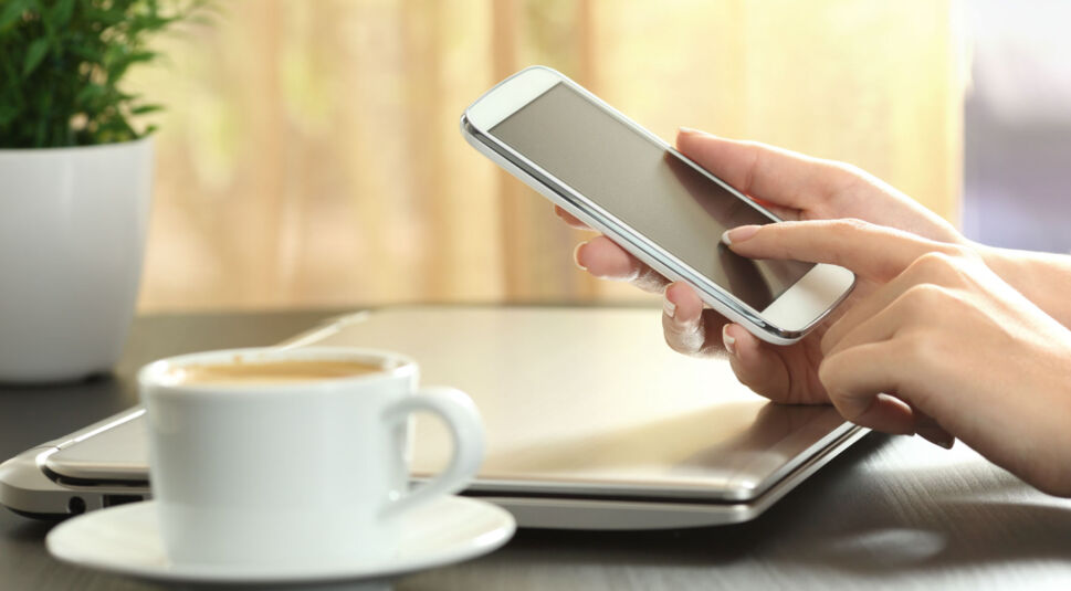 Frauenhände halten ein Smartphone und vor ihr steht Kaffee auf dem Tisch.