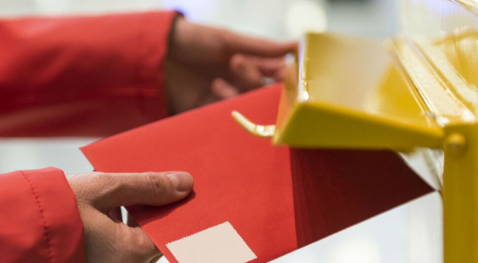 Roter Brief wird in gelben Briefkasten gesteckt.