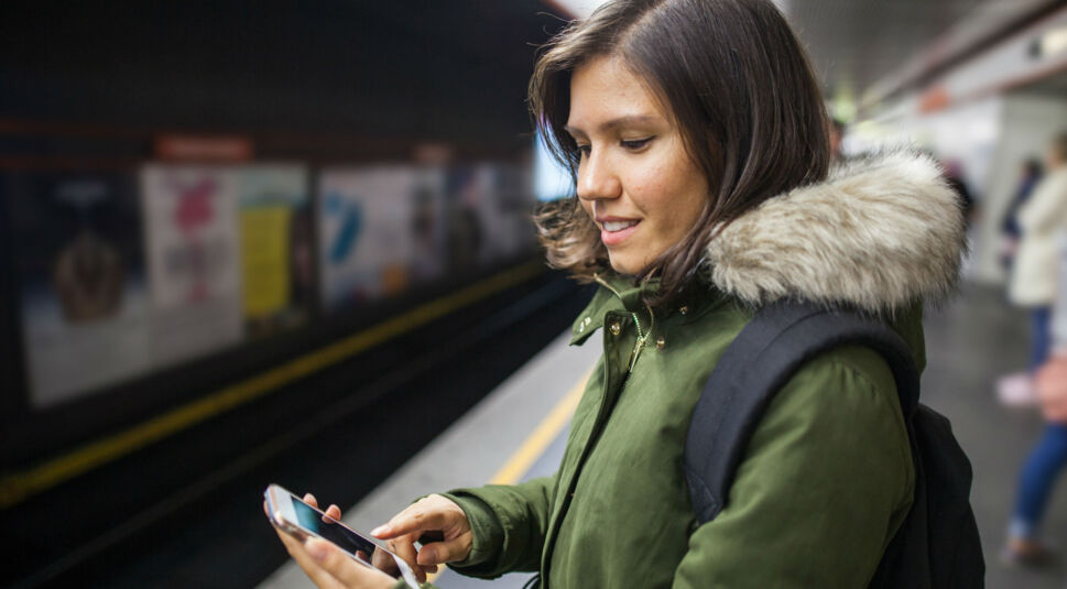 Lächelnde Frau am Bahnsteig schaut auf ihr Smartphone.