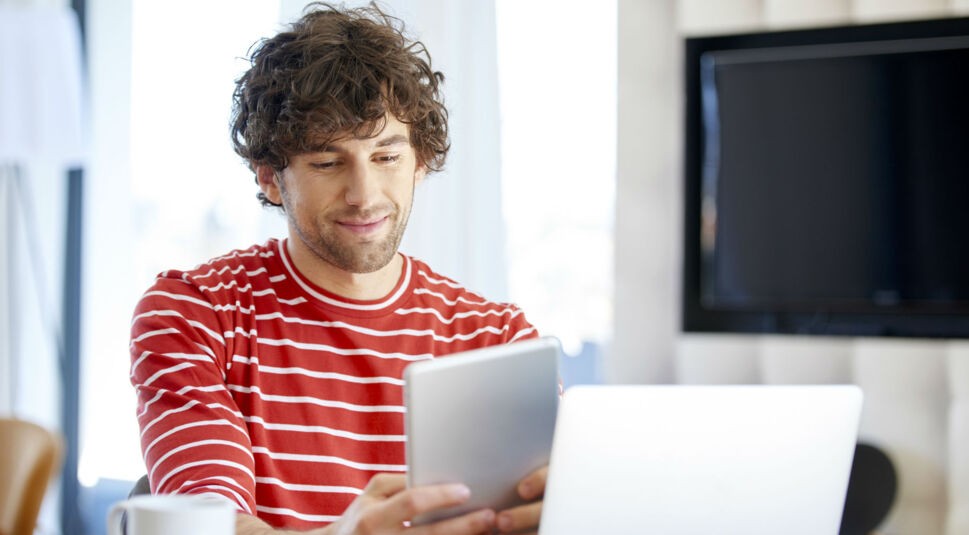 Mann sitzt am Tisch und liest am Tablet.