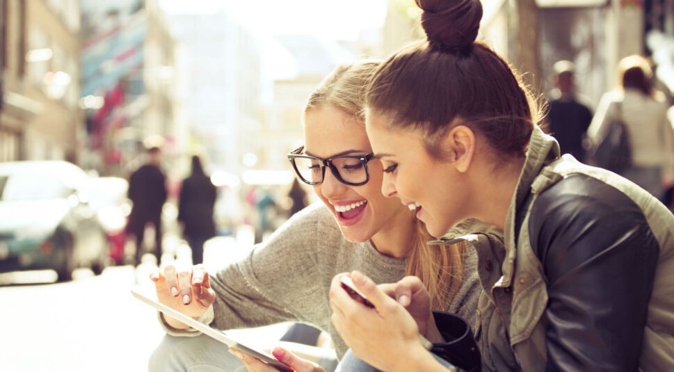 Zwei Freundinnen sitzen am Straßenrand und schauen auf ihr Tablet.