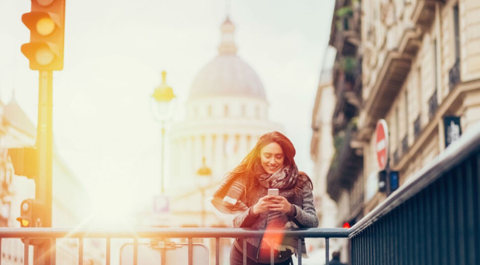 Junge Frau steht am Brückengeländer und schaut auf ihr Smartphone.