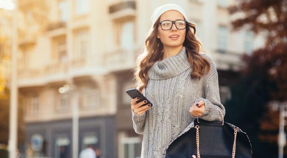 Junge Frau in grauem Pulli hält Smartphone in der Hand.