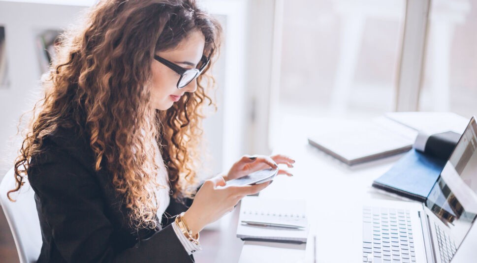 Junge Frau sitzt am Schreibtisch und spielt mit ihrem Smartphone.