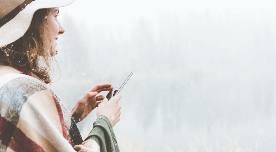 Junge Frau steht lächelnd im Schnee und hält dabei ein Smartphone in der Hand.