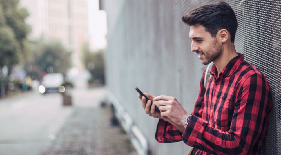 Junger Mann lehnt an Hauswand und tippt auf seinem Smartphone.