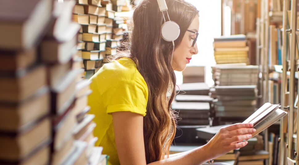 Eine junge Frau sitzt in der Bibliothek und hört ein Hörbuch während sie liest