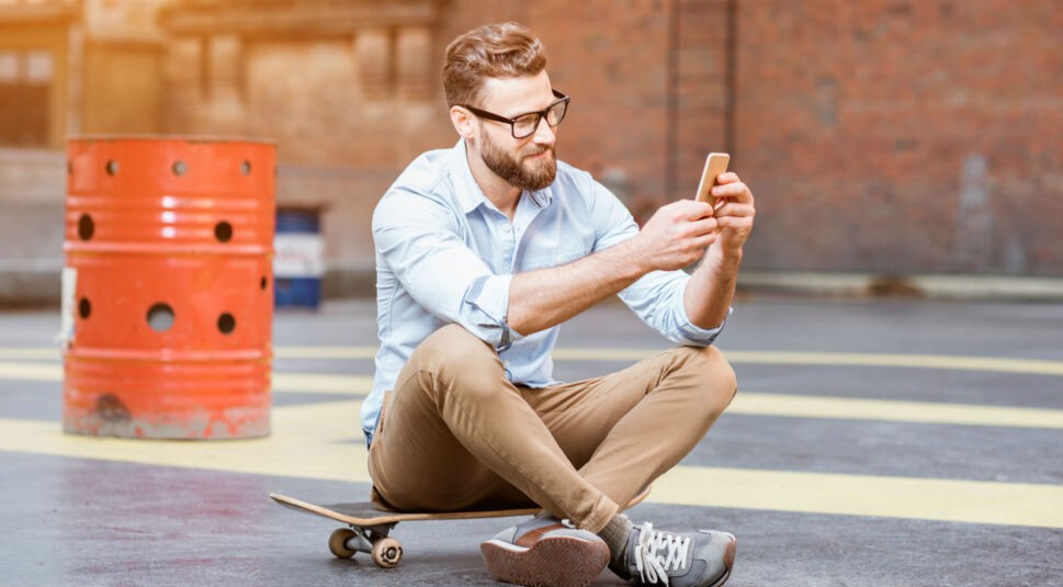 Junger Mann sitzt auf dem Skateboard und nutzt sein Smartphone.