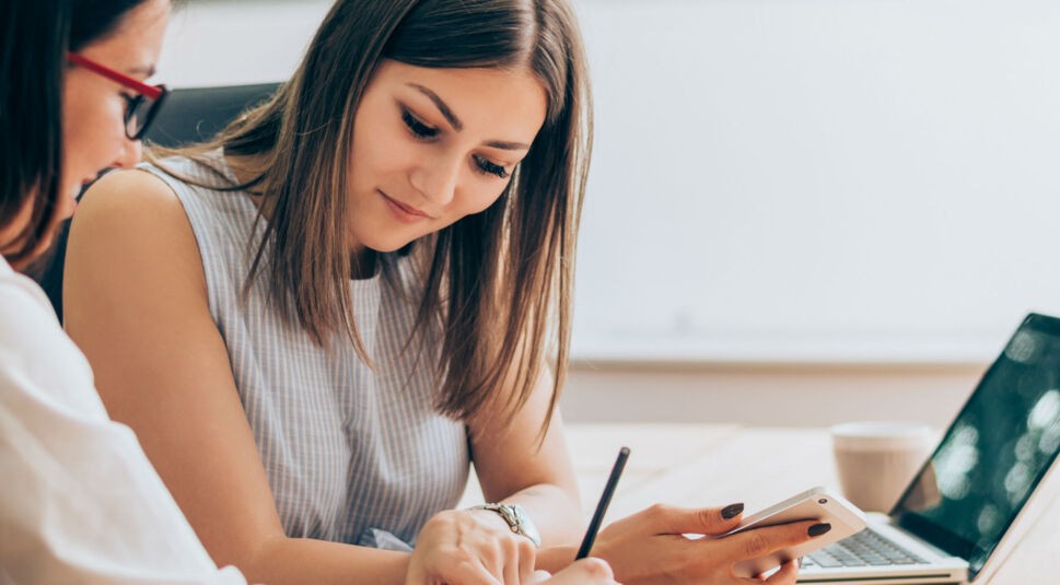 Junge Frau arbeitet mit ihrer Freundin am Laptop und hält ihr Smartphone in der Hand.