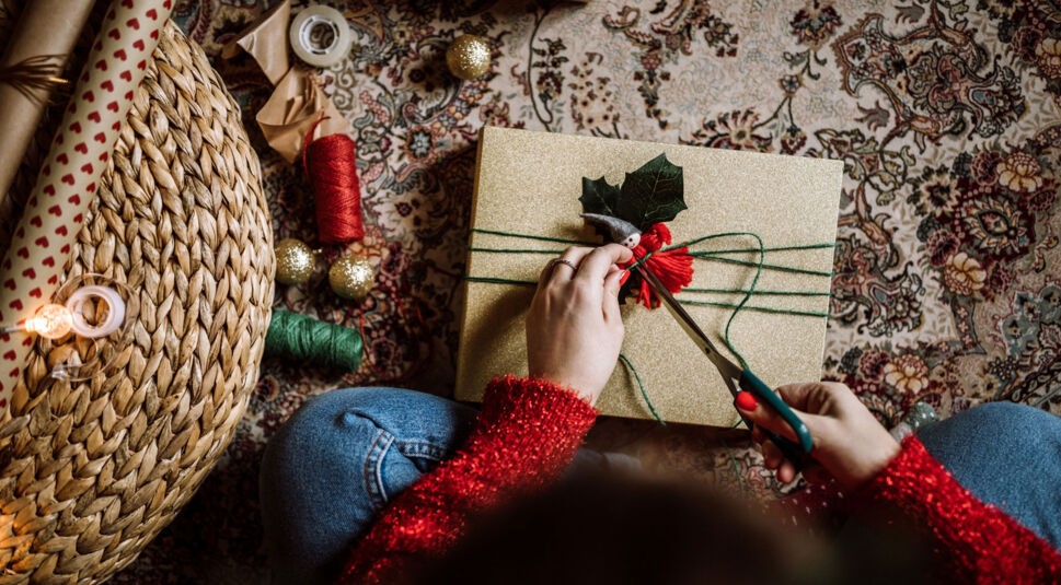 Eine Frau mit rotem Pulli packt ein Weihnachtsgeschenk ein und verziert es, neben ihr liegt Garn und Geschenkpapier..