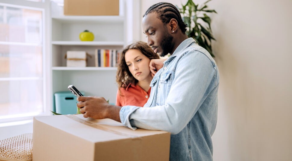 Auf dem Bild in dem Artikel zum Umzugsservice von Vodafone stehen ein junger Mann und eine Frau mit lockigem Haar vor einem großen Umzugskarton. Beide schauen konzentriert auf das Smartphone des Mannes.
