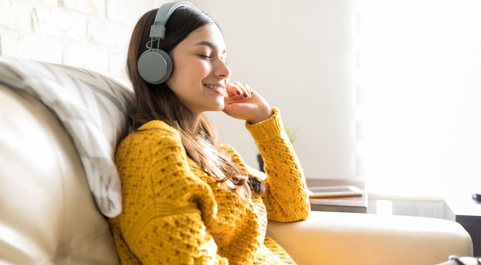 Frau mit Kopfhörern und gelben Pulli hört zufrieden Mindfulness-Podcasts auf dem Sofa