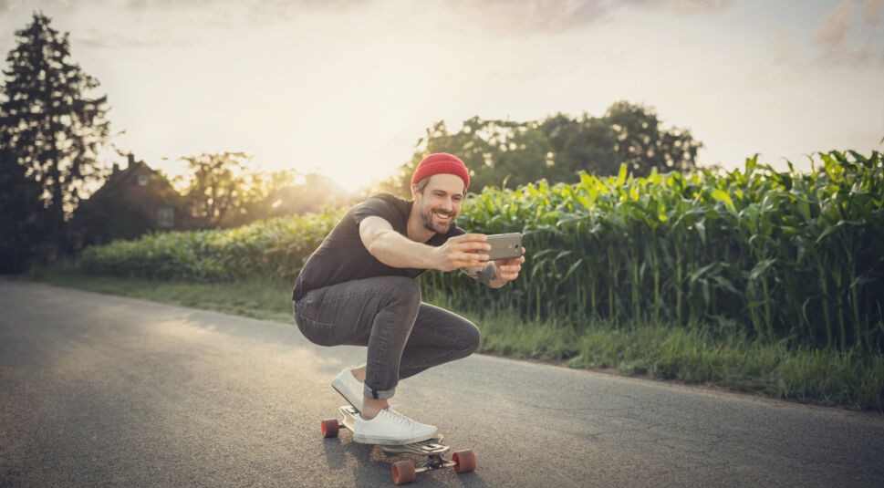 Ein Mann fährt mit seinem Skateboard an einem Feld vorbei und nimmt sich dabei auf.