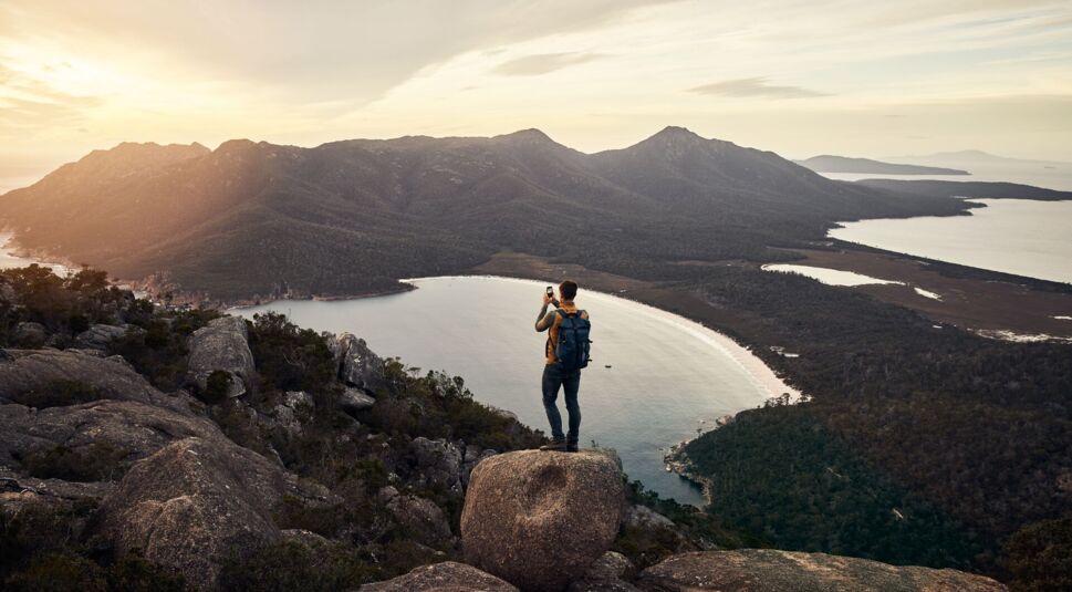 Ein Mann mit Wanderausrüstung steht auf einem Bergfels und macht ein Foto vom See, der darunter liegt.