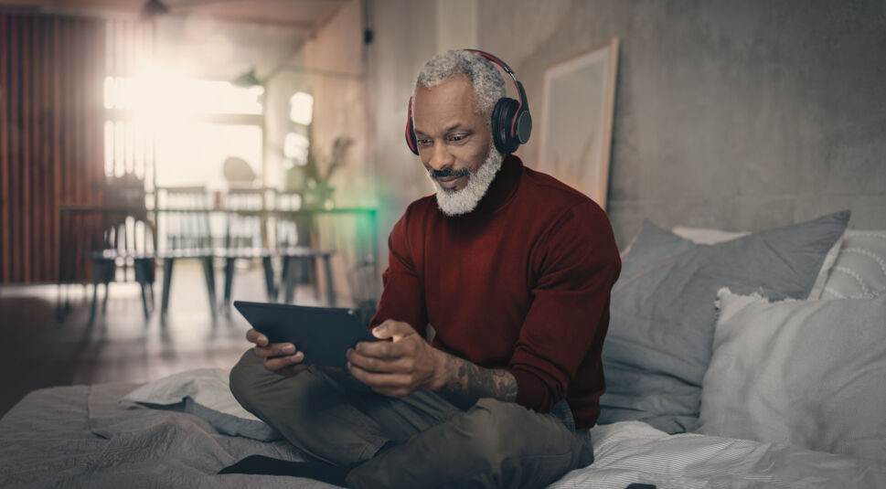 Ein Mann sitzt mit Kopfhörern und seinem Tablet auf dem Bett.