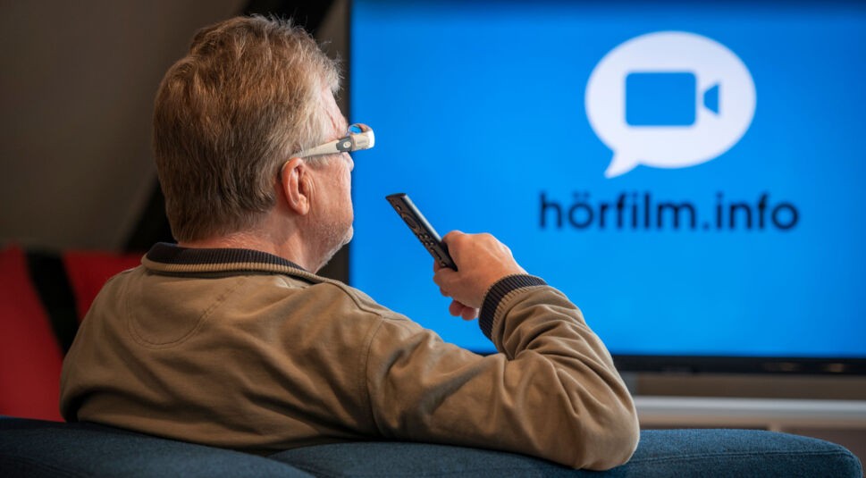 Ein Mann hört sich über den Alexa-Hörfilm-Skill einen Inhalt mit Audiodeskription an, der TV-Bildschirm ist eingeschaltet.