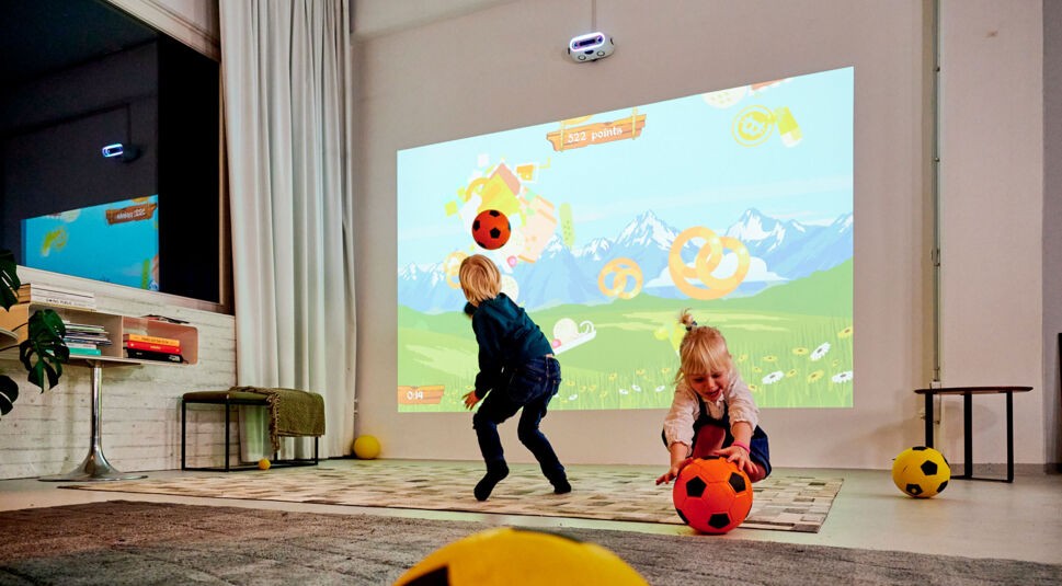 Zwei Kinder spielen mit Bällen, die sie gegen eine Wand werfen, auf die ein Spiel projiziert ist.