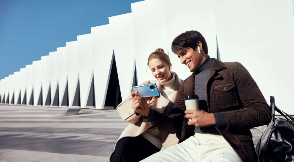 Ein Mann zeigt einer Frau eine Inhalt auf seinem Xiaomi Mi 11 | 5G, er trägt Kopfhörer und hält einen Kaffeebecher.