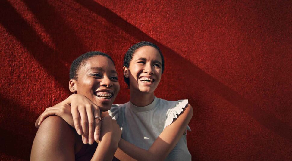 Zwei Mädchen stehen lachend vor einer roten Wand, die rechte umarmt die linke.