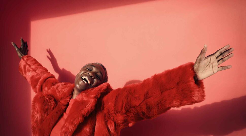 Eine Frau mit rotem Fake-Fur-Mantel streckt lachend die Arme aus.