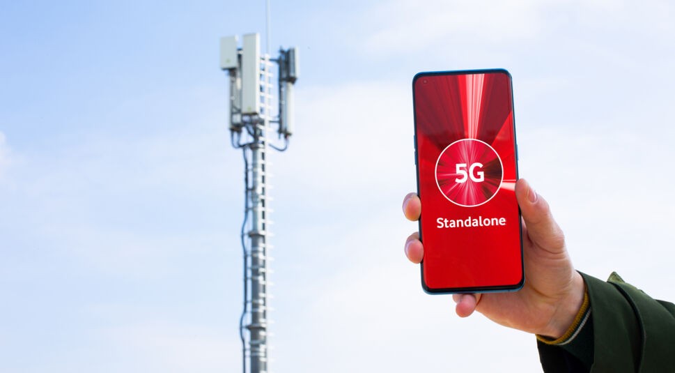 Ein Smartphone, das "5G Standalone" zeigt, dahinter ein Mobilfunkmast.