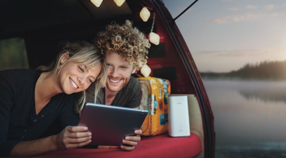 Ein Paar liegt im Kofferraum seines Campers und schaut sich etwas auf dem Tablet an.