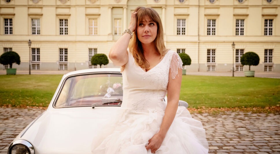 Eine junge Frau in einem Hochzeitskleid sitzt auf der Haube eines Autos