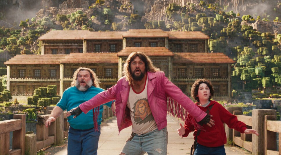 In dieser Szene aus dem Minecraft-Film ist Jason Momoa in einer auffälligen pinken Lederjacke zu sehen, wie er zusammen mit zwei weiteren Charakteren erschrocken nach vorne blickt. Die Kulisse zeigt eine typische Blockwelt aus dem Spiel Minecraft mit pixeligen Bäumen und Gebäuden. Ideal für Fans von Jason Momoas beste Filme und Serien.