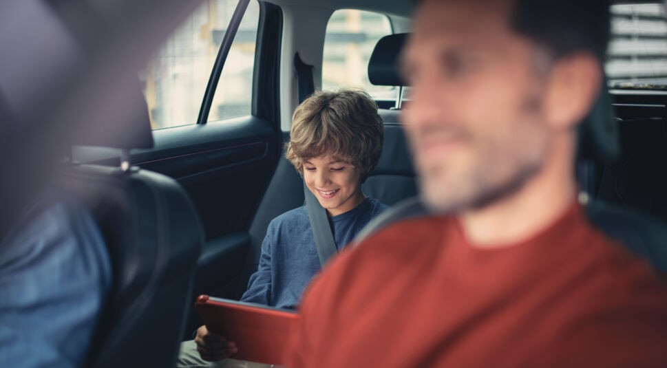 Eine Familie fährt im Auto, der Sohn sitzt auf dem Rücksitz liest etwas auf einem Tablet.