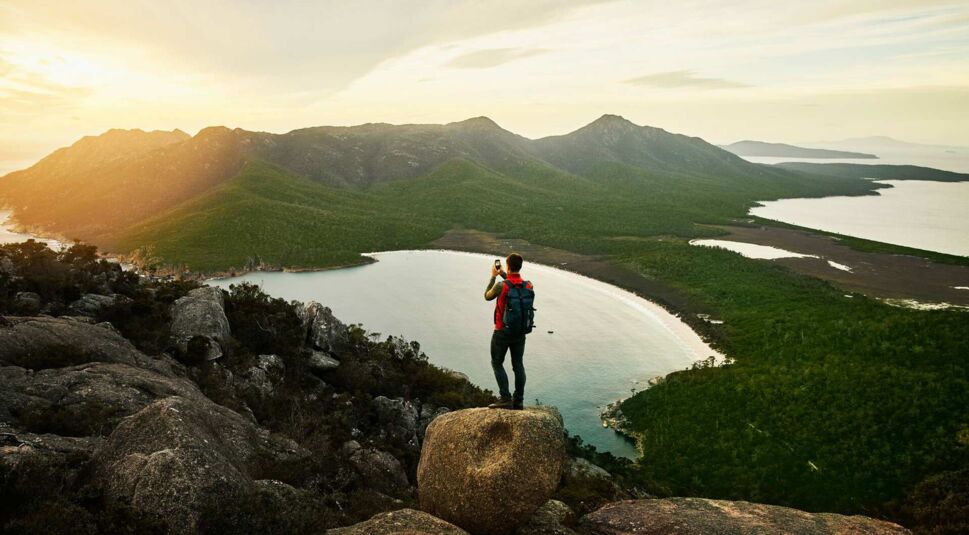 Ein Mann steh auf einem Berg und fotografiert den See im Tal mit seinem Smartphone.