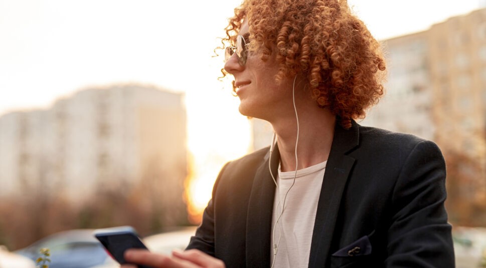 Ein Mann mit Sonnenbrille ist draußen unterwegs und hört per Kopfhörer etwas auf seinem Handy an. 