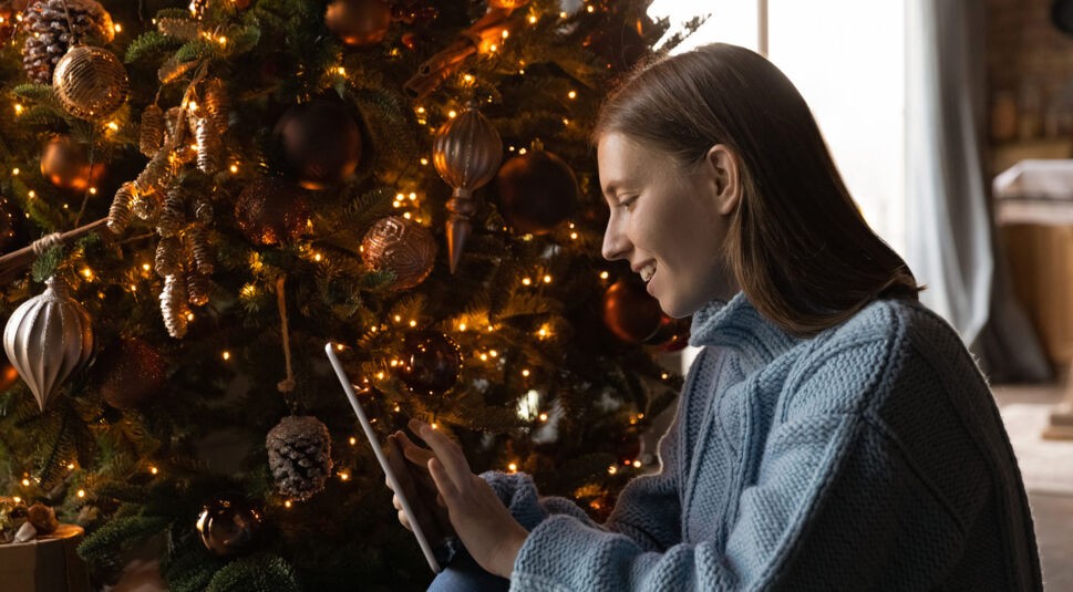 Eine Frau sitzt vor dem Weihnachtsbaum und scrollt auf ihrem Tablet.