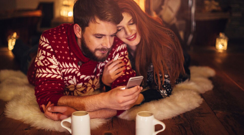 Ein Paar liegt in Weihnachts-Pullovern auf dem Boden und schaut sich etwas auf dem Smartphone an.