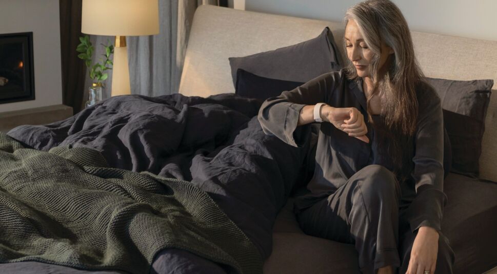 Ältere Frau sitzt im Bett und guckt auf ihre Smartwatch Fitbit Sense