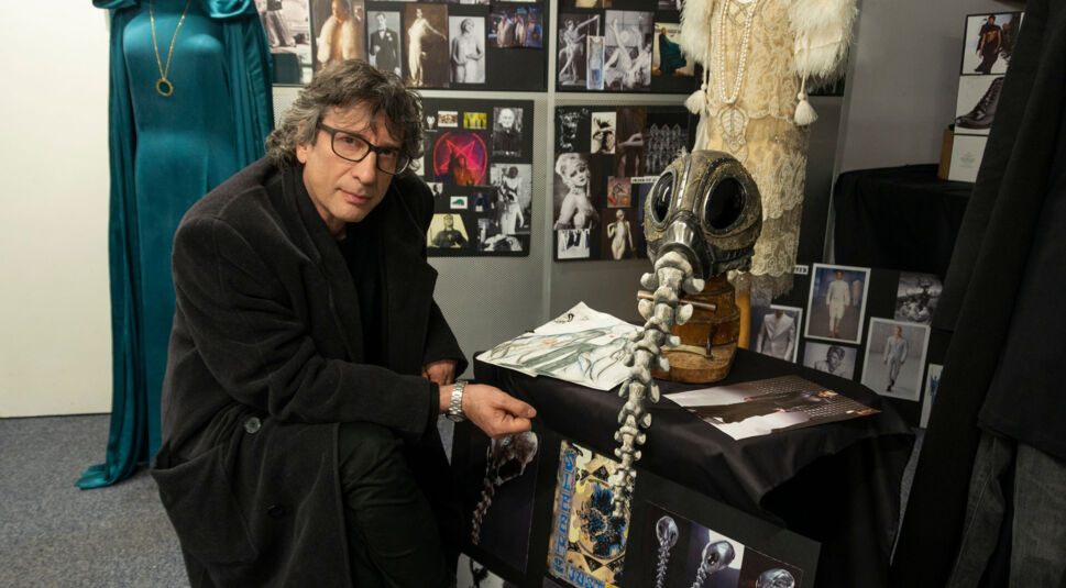 Neil Gaiman sitzt neben dem Helm vom Sandman und schaut in die Kamera