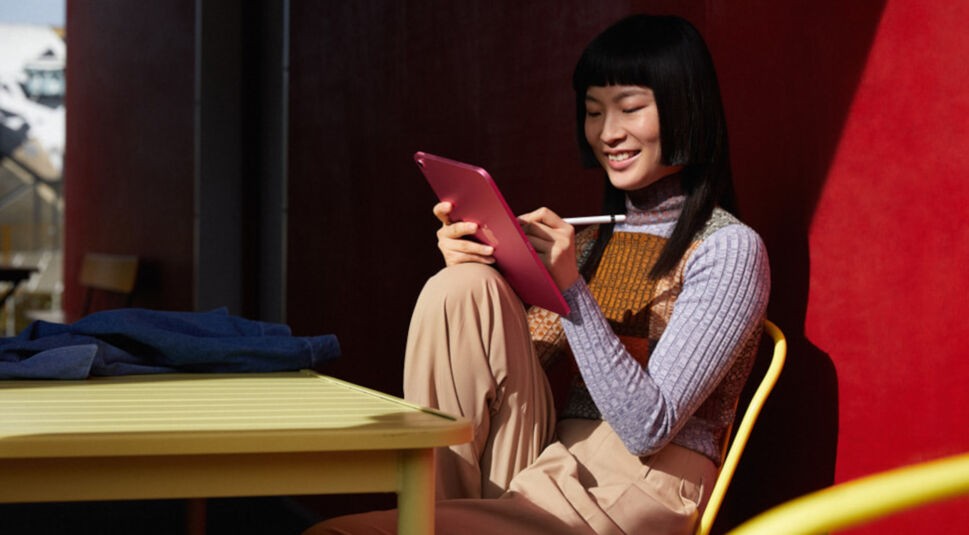 Schwarzhaarige mit pinkem Apple iPad 10 und Apple Pencil arbeitet entspannt an eine Hauswand gelehnt