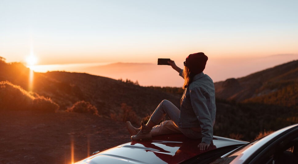 Frau sitzt auf Auto und fotografiert den Sonnenuntergang / Naturfotos mit dem iPhone 14