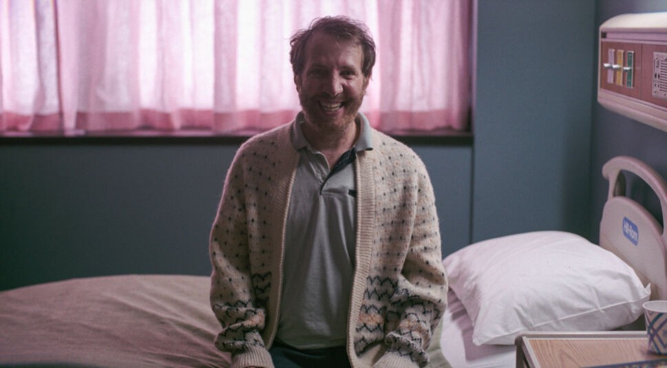 Ein Mann sitzt auf einem Krankenhausbett und grinst grotesk