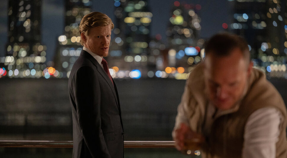 Auf dem Bild zur Serie Zero Day mit Robert De Niro ist Jesse Plemons in einem dunklen Anzug mit roter Krawatte zu sehen. Er steht auf einer Terrasse bei Nacht und blickt ernsthaft über die Schulter, während die Lichter einer Stadt verschwommen im Hintergrund funkeln. Im Vordergrund ist eine unscharfe Person zu erkennen, die ein Getränk hält, was der Szene eine geheimnisvolle und angespannte Atmosphäre verleiht.